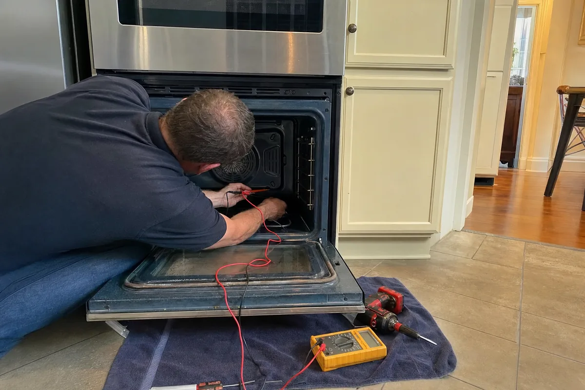 A technician reaching deep into a built-in oven cavity with meter leads run down to the clamp meter on the floor