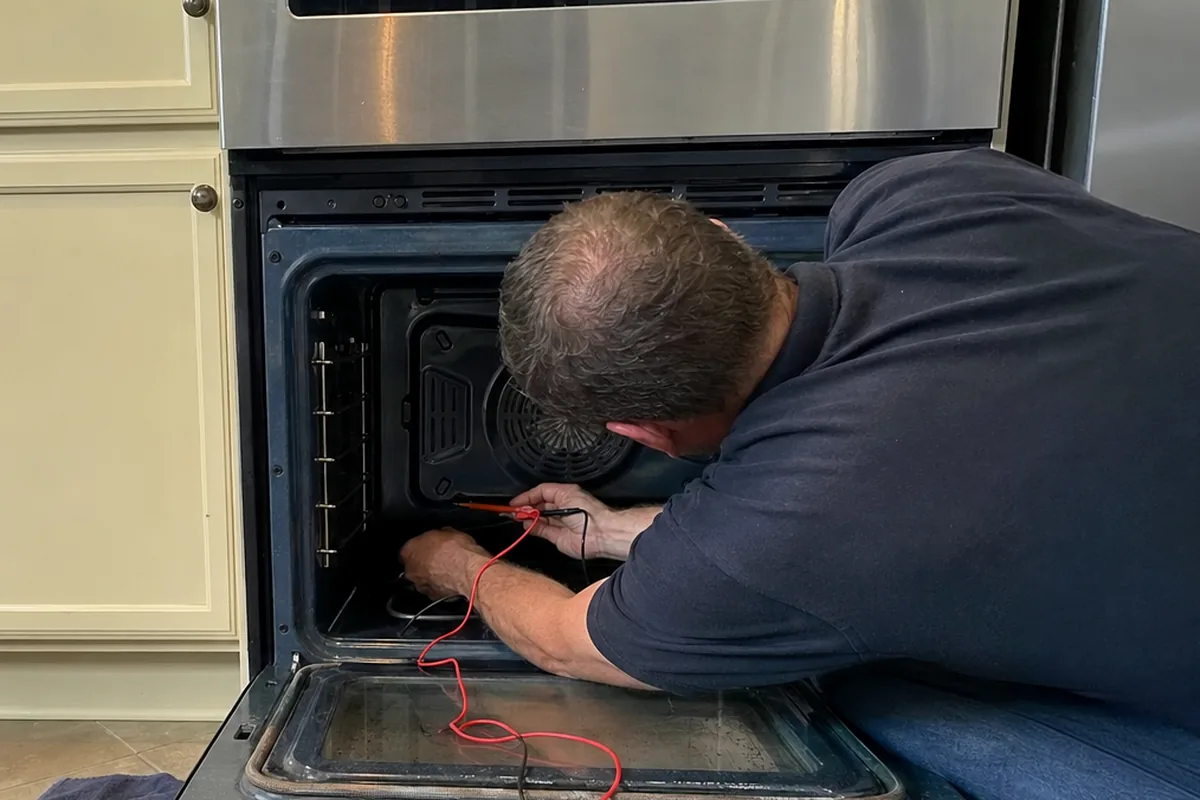 A technician at an open built-in oven with the meter on the floor mat beside his knees