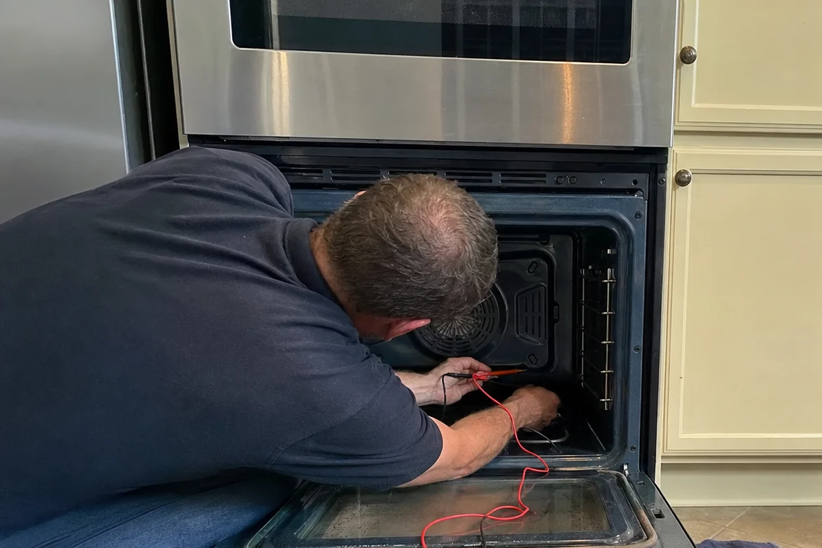 A technician kneeling right inside a built-in oven, red meter leads trailing over the dropped door towards the fan cover at the back of the cavity