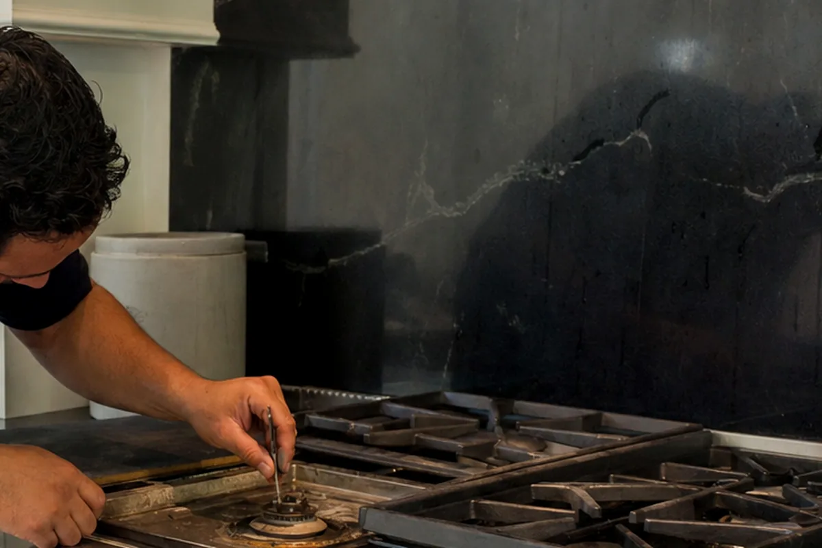 A range burner being worked on with the grates off, daylight from a window falling across the stone counter