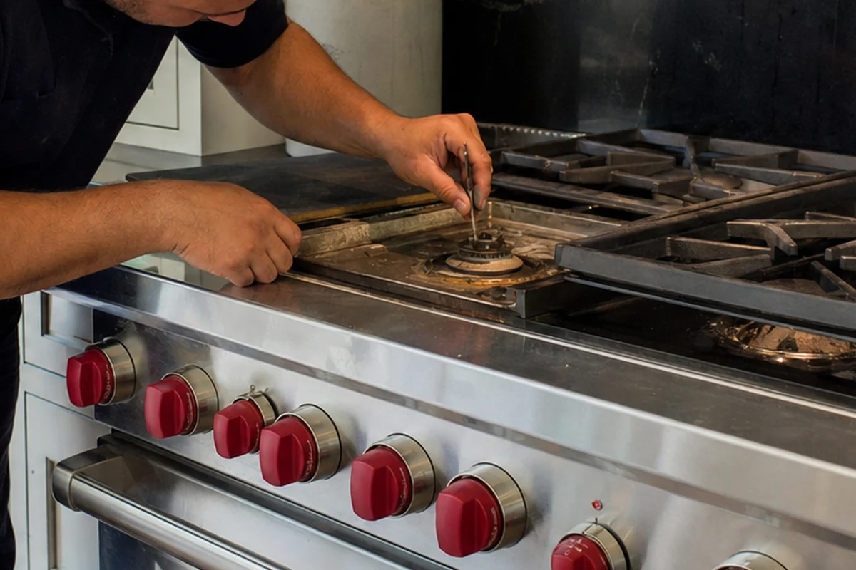 Both hands working at an uncovered burner well on the deck of a range, the row of red control knobs running along the front below